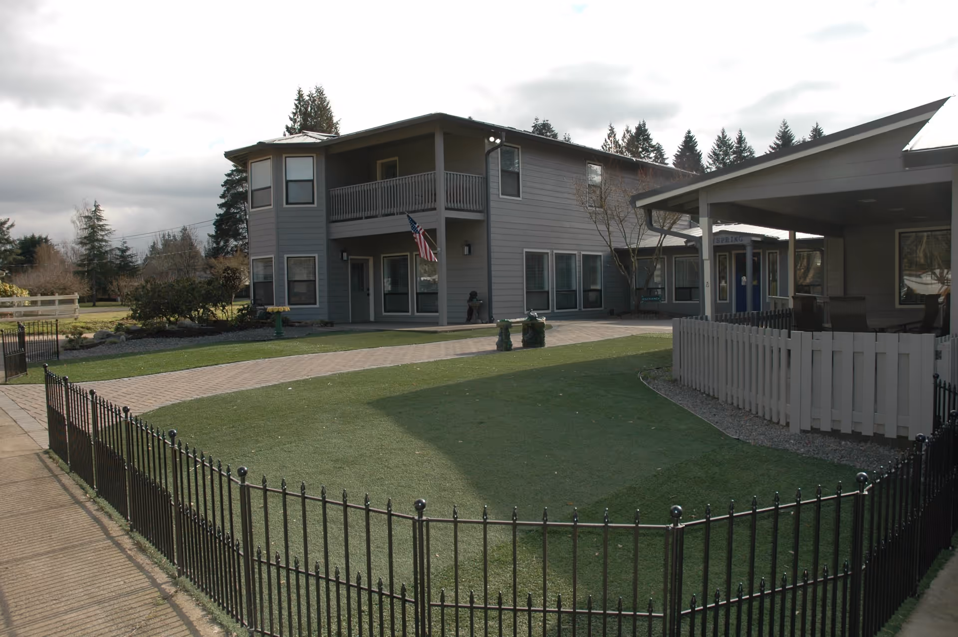 Exterior view of Dayspring Memory Care facility showing a two-story gray building with multiple windows and a balcony. There is a paved walkway and a fenced grassy area in the foreground. An American flag is displayed near the entrance, and trees are visible in the background under a cloudy sky.