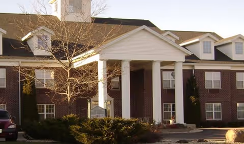 Exterior view of a two-story brick building with white columns at the entrance and multiple windows. There are some bushes and a leafless tree in front of the building under a clear sky.