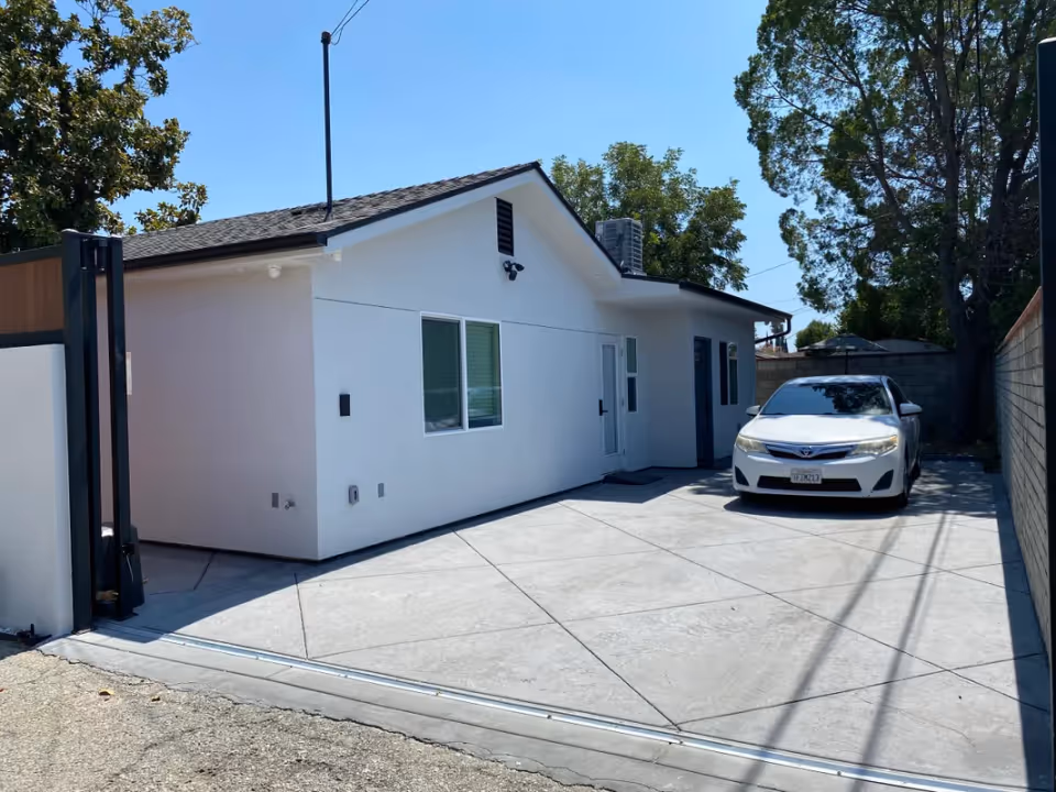 Driveway and entrance of a small white single-story building with a parked white car and trees in the background.