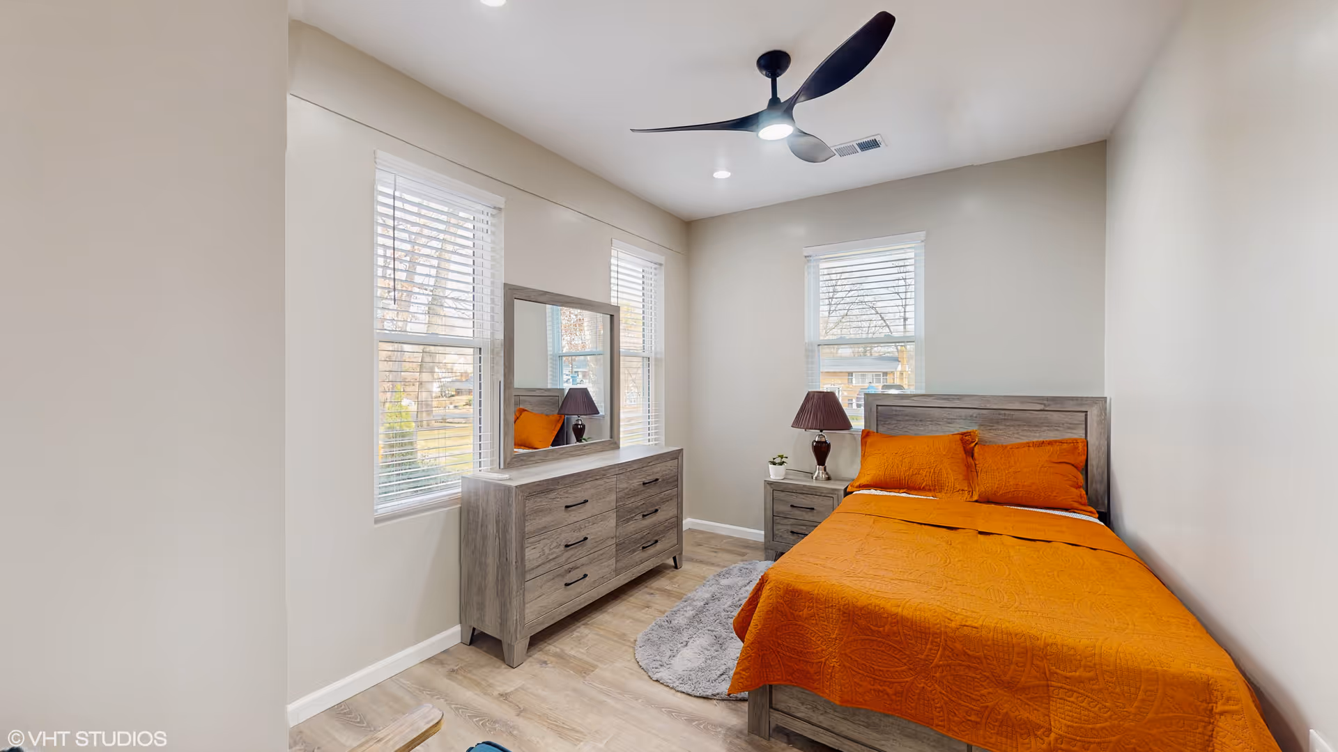 A furnished bedroom with orange bedding, gray wooden dresser, two windows, and ceiling fan.