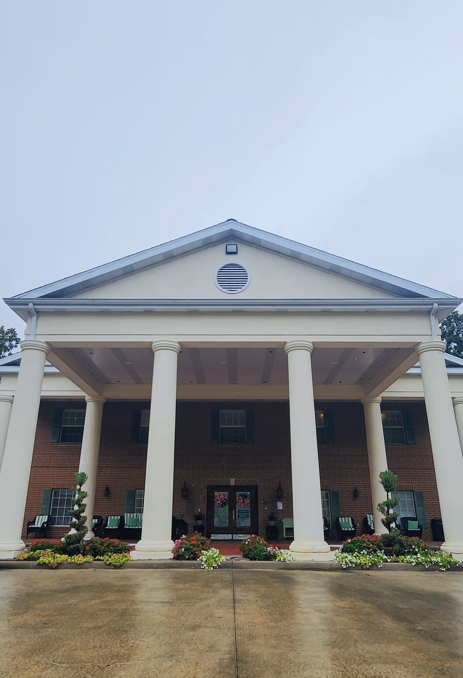 Front view of a brick senior living facility with a columned porch and flower beds.