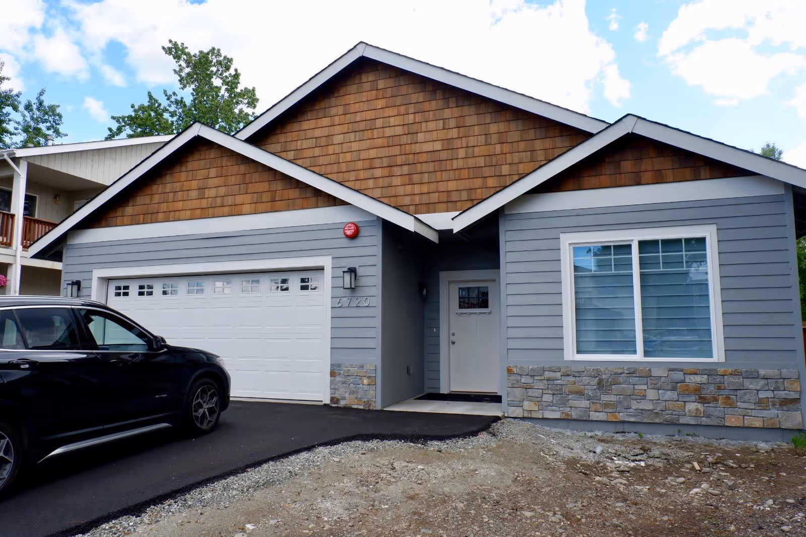 Front exterior view of a single-story house with gray siding, stone accents, and brown shingle gables. There is a white garage door, a white front door with a small window, and a black car parked in the driveway. The sky is partly cloudy.