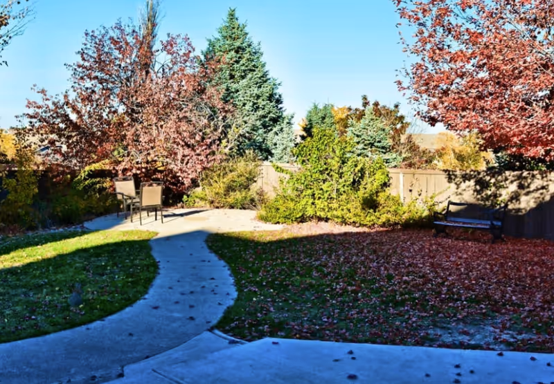 Sunny outdoor courtyard with curved walkway, benches, chairs, and colorful autumn trees.