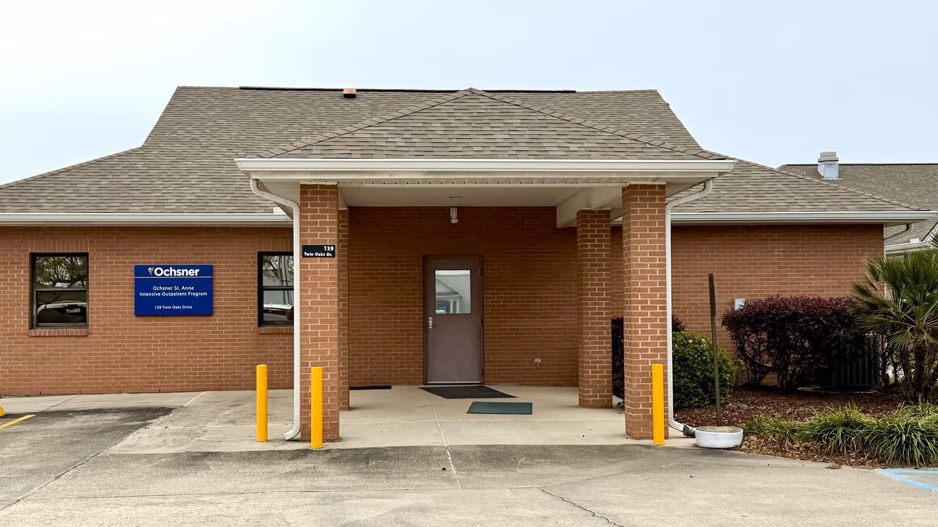 Brick building entrance with a blue Ochsner St. Anne Intensive Outpatient Program sign and yellow bollards.