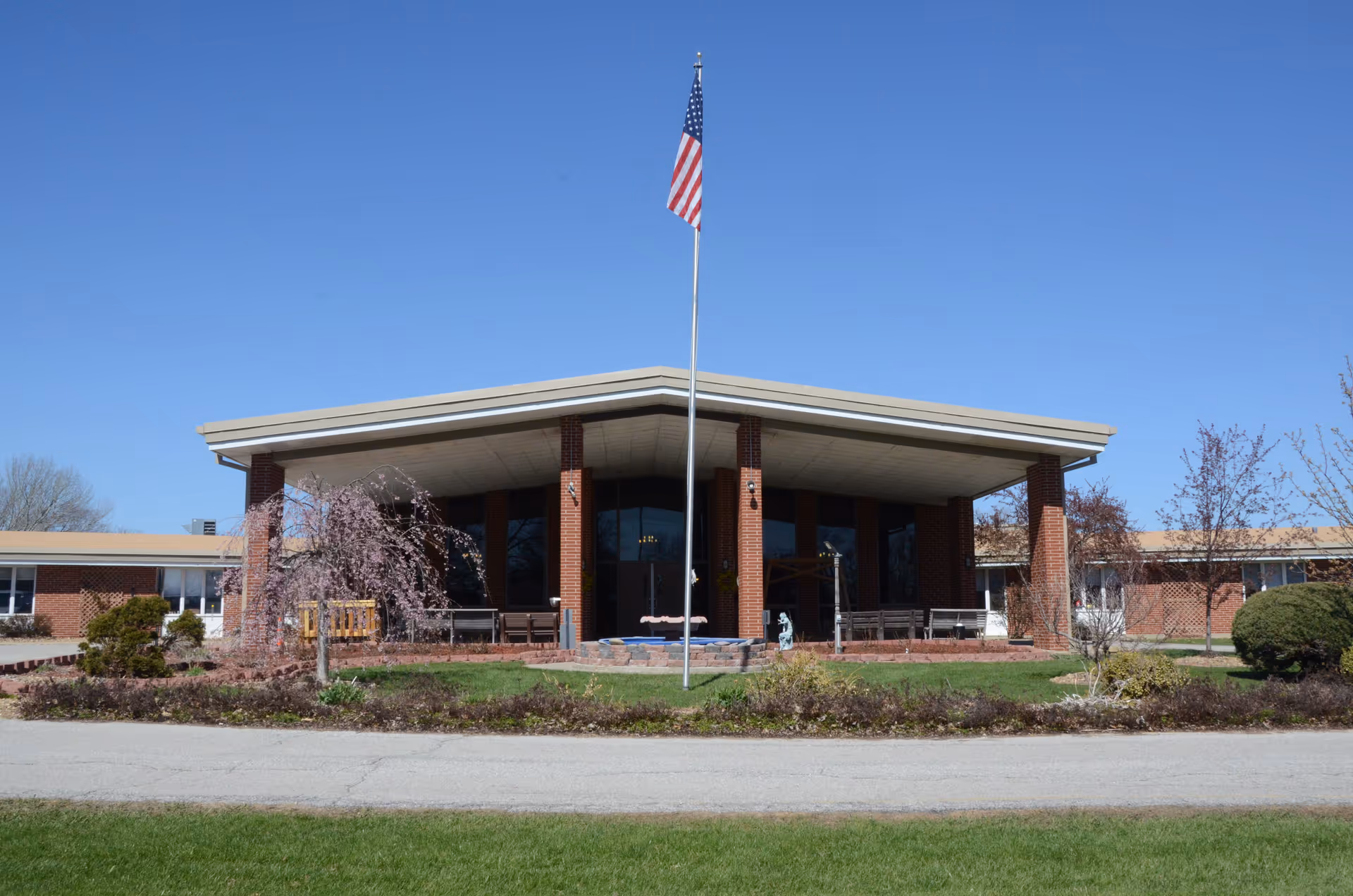 Brick senior living facility entrance with flagpole, benches, and landscaped lawn.