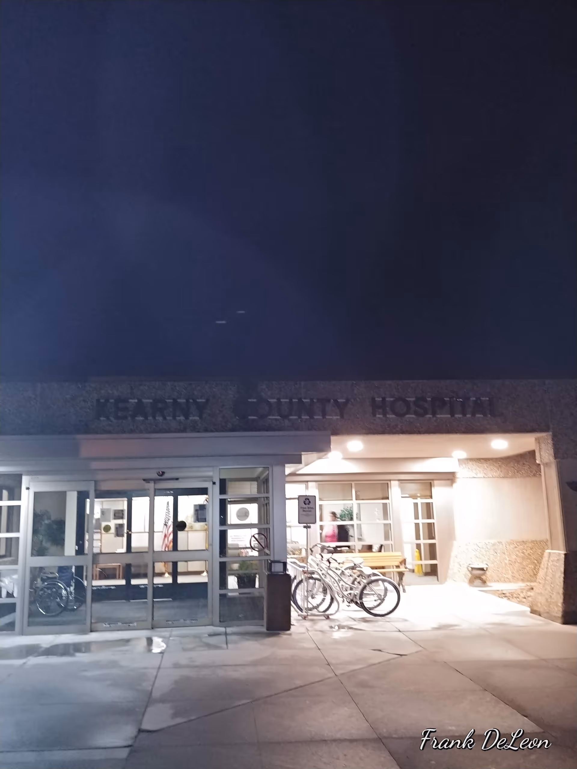 Night exterior of Kearny County Hospital entrance with glass doors and bicycles parked outside.
