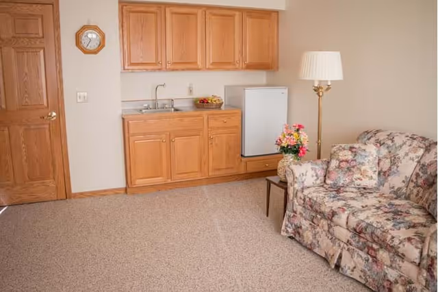 A small living area with a floral patterned couch and matching pillow, a wooden side table with a vase of flowers, a tall floor lamp, and a kitchenette with wooden cabinets, a small sink, and a mini refrigerator. There is a wooden door and a wall clock above the light switch.