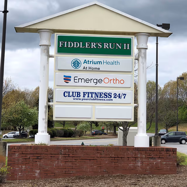 Exterior signboard on a brick base listing Atrium Health At Home among other businesses.