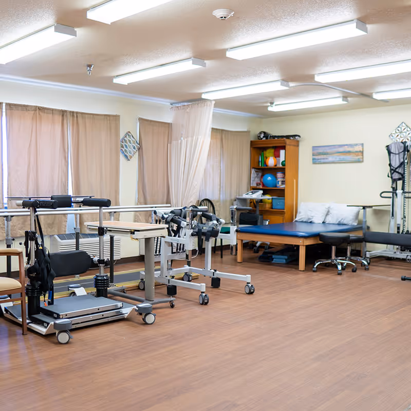 An indoor physical therapy room with parallel bars, exercise equipment, a treatment table, and storage shelves.