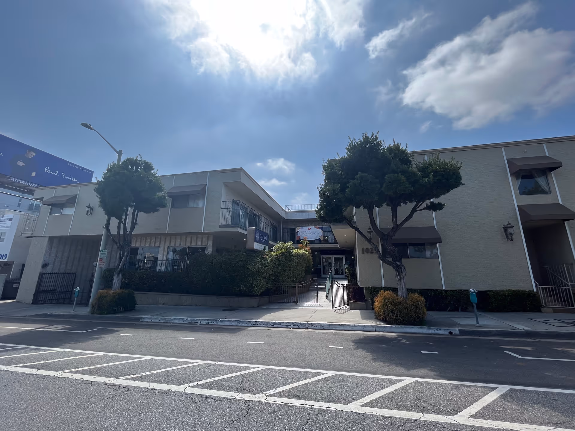 Front exterior of a two-story senior living building with trees, sidewalk and entrance under a bright sky.