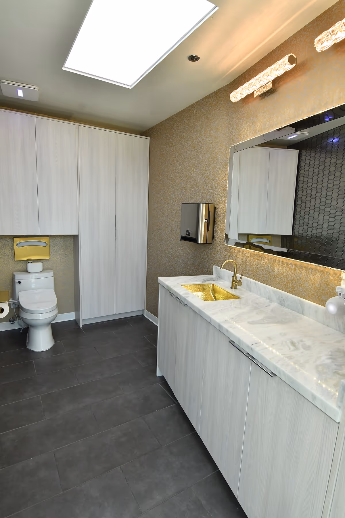 Modern public bathroom with gray cabinetry, marble countertop, gold sink, and toilet.