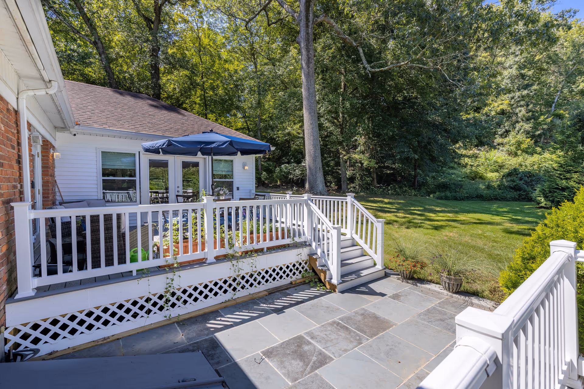 White deck with railing, umbrella, and seating overlooks a green backyard and trees.
