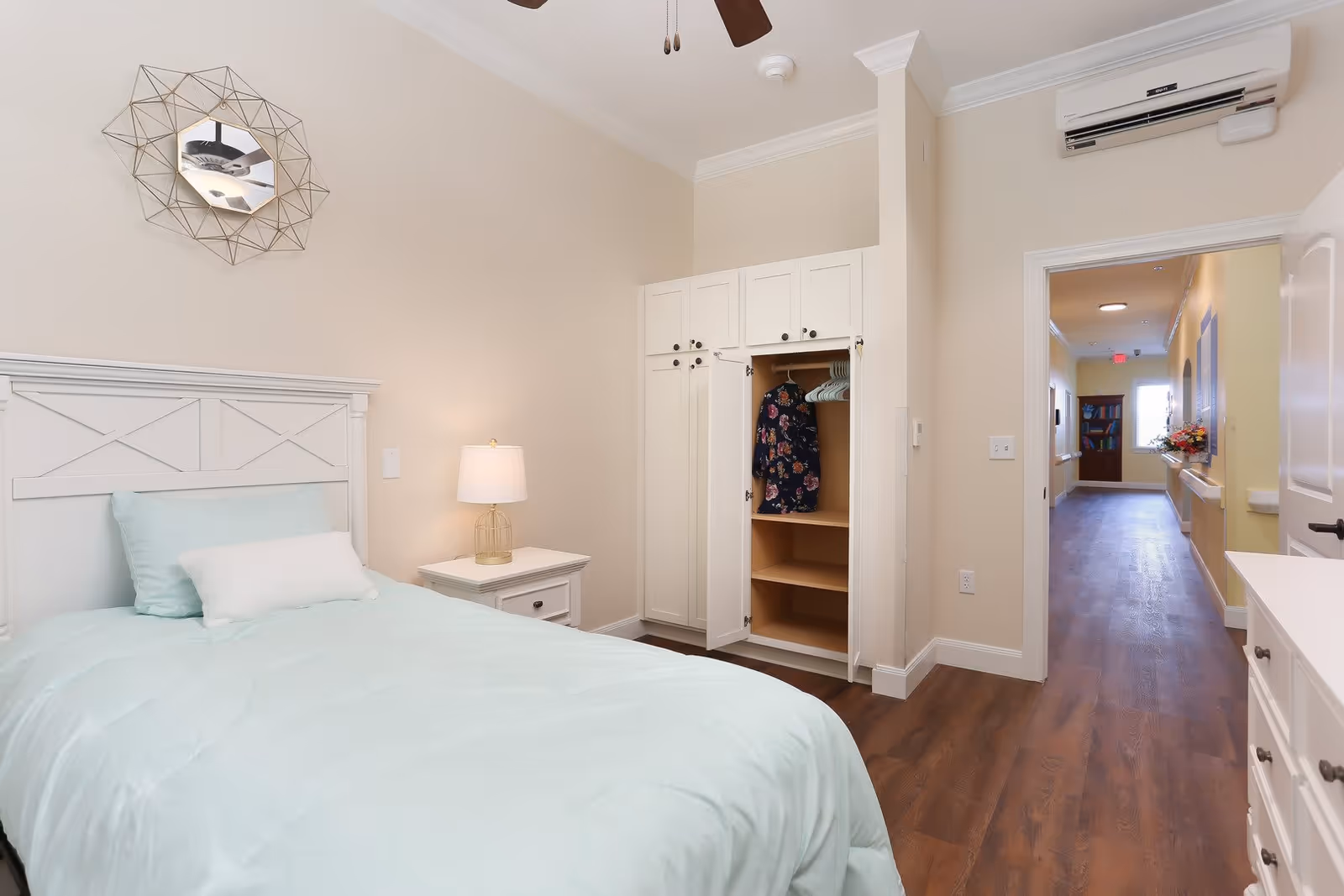 A bright and clean bedroom in an assisted living facility featuring a single bed with light blue bedding and pillows, a white nightstand with a lamp, an open white wardrobe with a floral dress hanging inside, a decorative wall mirror above the bed, and a hallway visible through an open door.