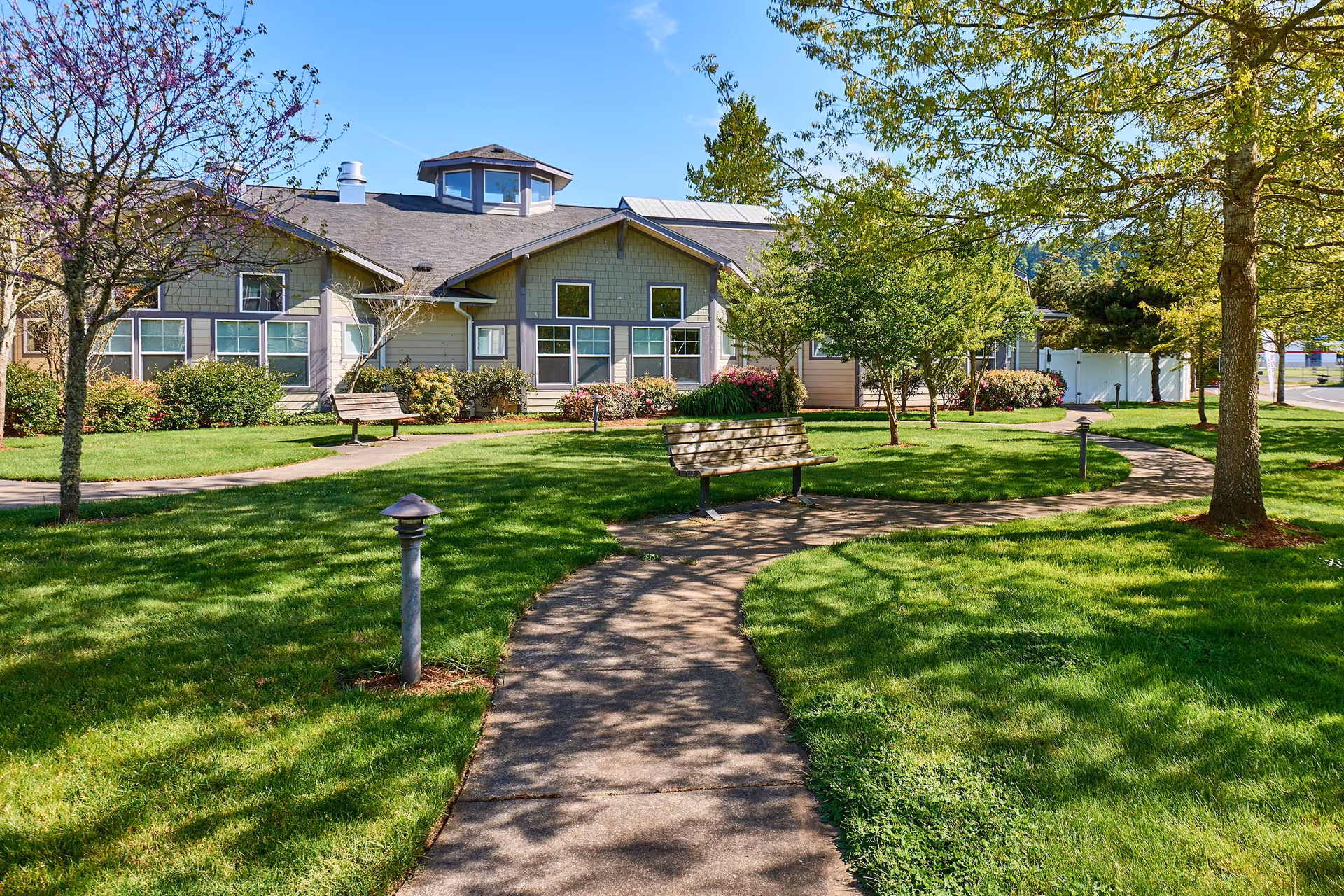 Landscaped front yard with a winding sidewalk, benches, trees, and the exterior of a single-story assisted living building under a blue sky.