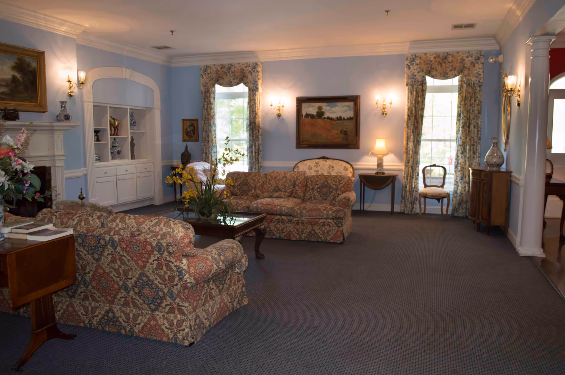 A cozy living room with patterned upholstered sofas and armchairs arranged around a wooden coffee table with a floral centerpiece. The walls are painted light blue and decorated with framed paintings and floral curtains on tall windows. There is a white fireplace on the left with decorative items on the mantel, and a built-in white shelving unit beside it. The room is softly lit by wall sconces and a table lamp on a small side table.