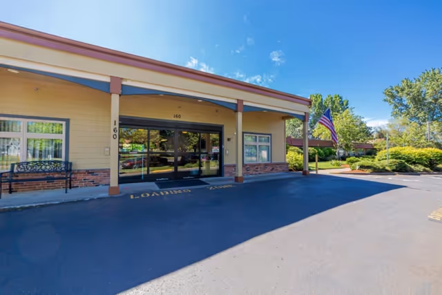 Entrance facade of Corvallis Manor Nursing & Rehabilitation Center with a covered drop-off area, glass doors, and an American flag.