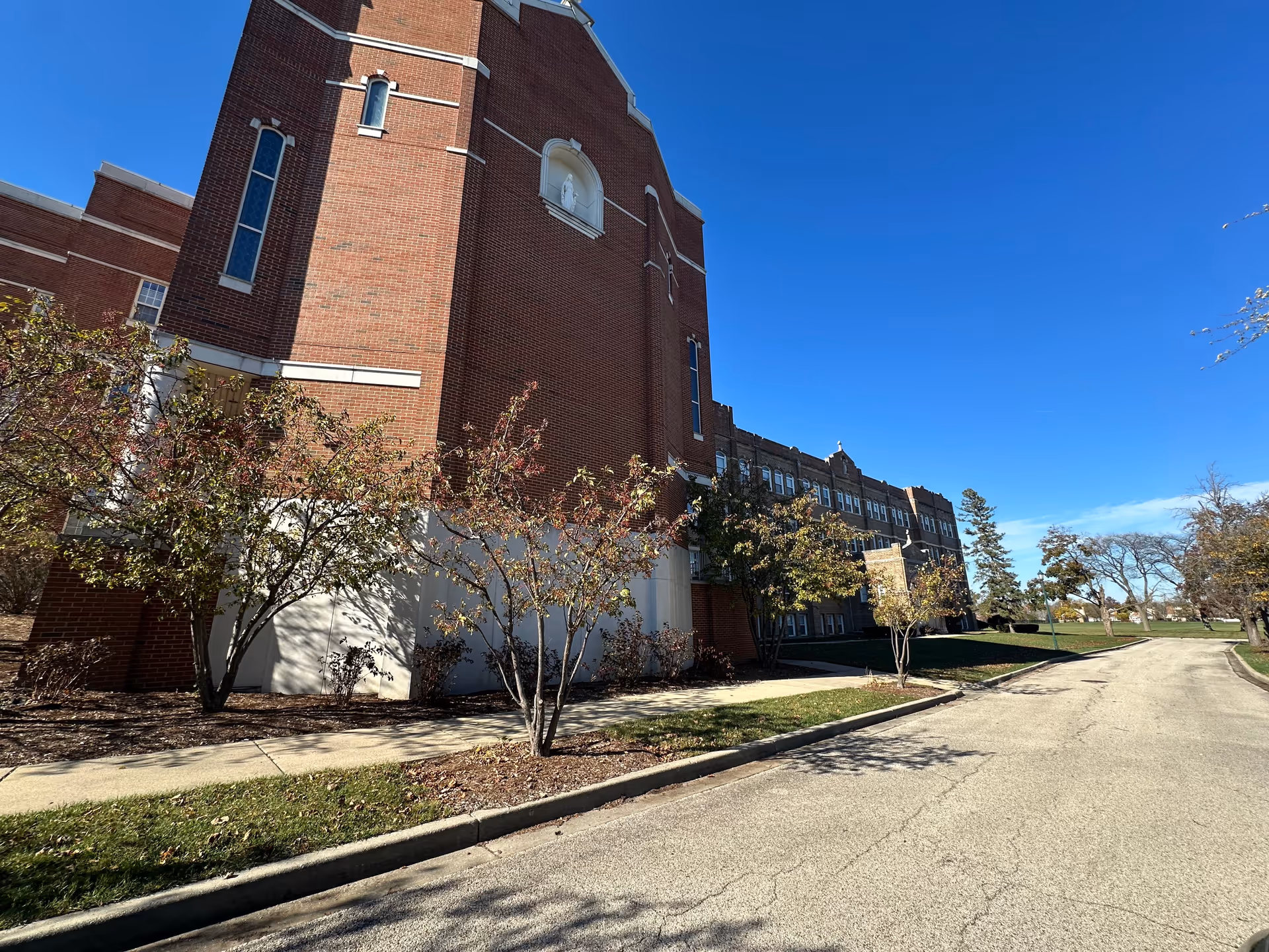 Exterior view of a large brick building with tall windows and a statue niche, surrounded by small trees and a sidewalk under a clear blue sky.