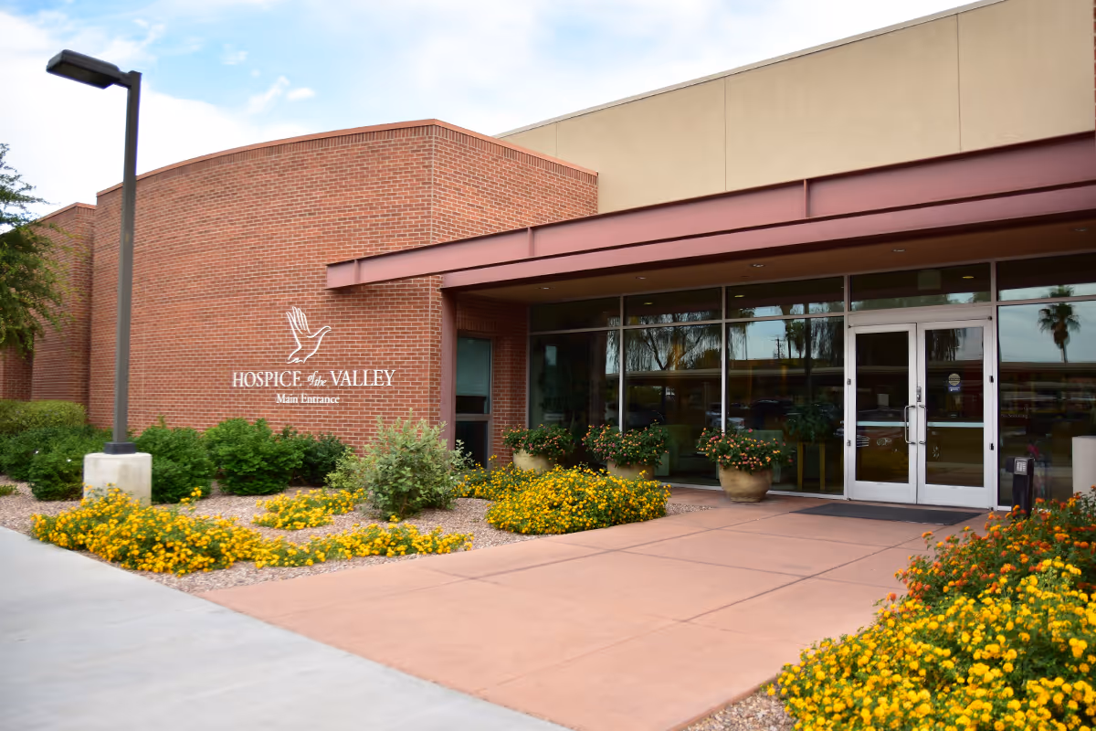 Brick building exterior with Hospice of the Valley main entrance and glass doors, surrounded by flowers.