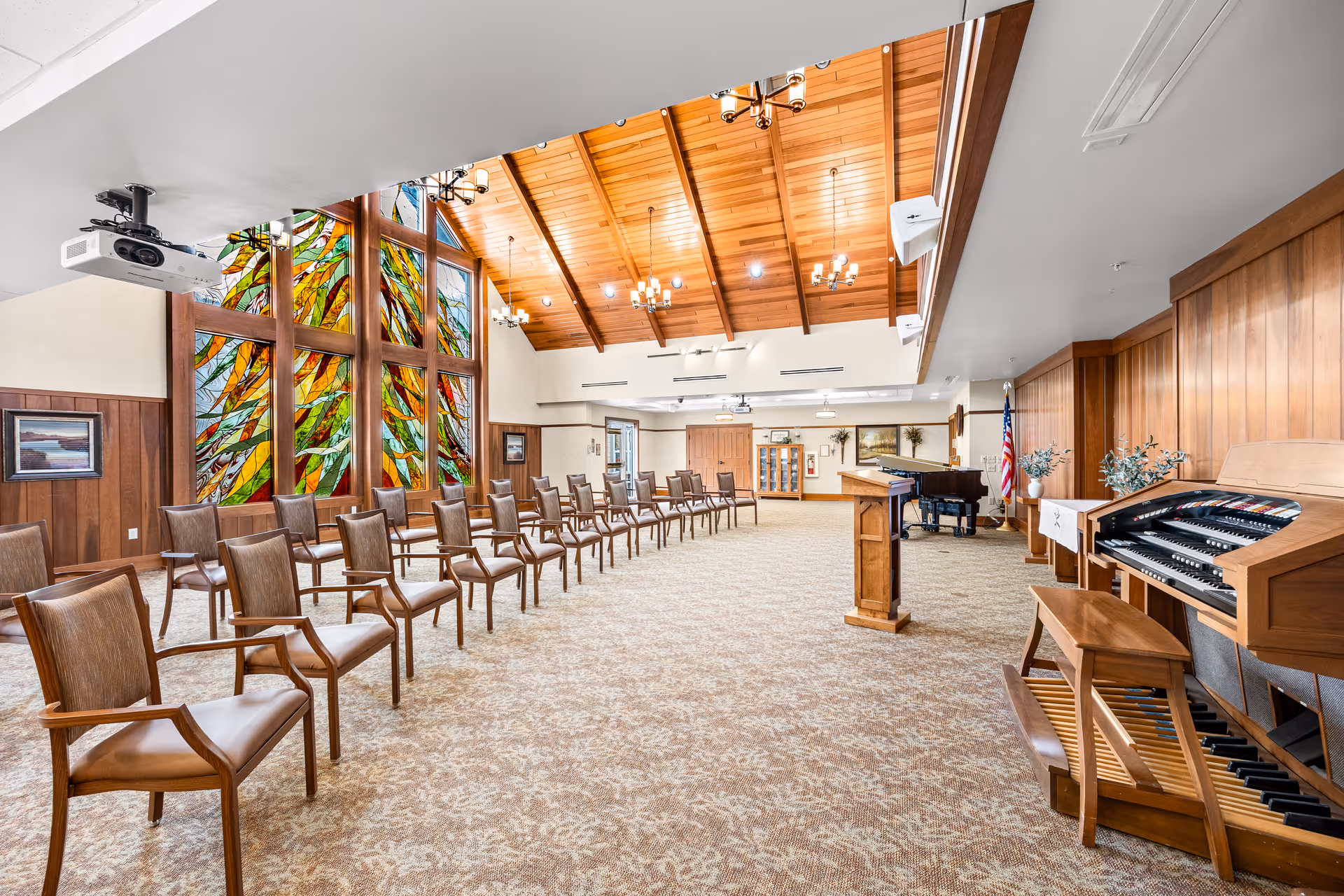 A spacious room with a high wooden vaulted ceiling and large colorful stained glass windows. The room has rows of wooden chairs with armrests arranged facing a wooden podium and a grand piano. There is also an organ with a bench on the right side. The walls are partly wood-paneled, and there are chandeliers hanging from the ceiling. An American flag stands near the piano.