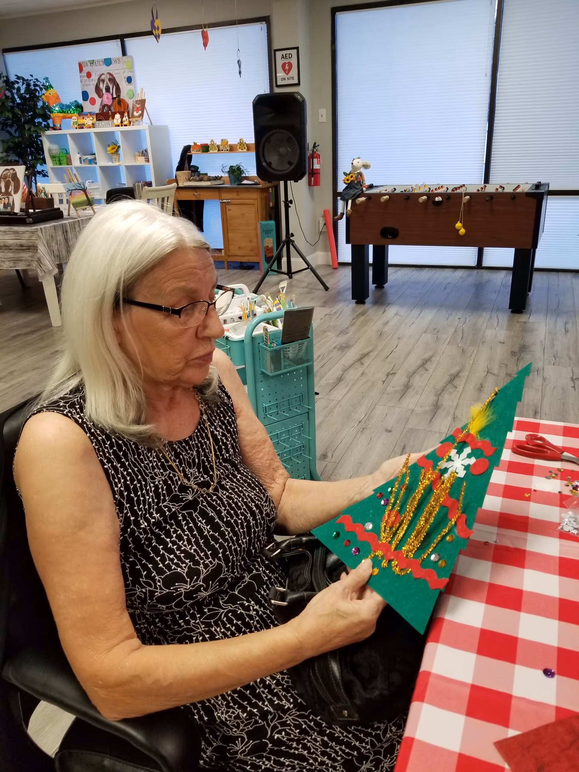 An elderly woman with white hair and glasses is sitting at a table covered with a red and white checkered tablecloth, holding a green triangular craft decorated with glitter, sequins, and other colorful embellishments. The room has wooden flooring and is decorated with various items including a foosball table, a speaker, a fire extinguisher, and shelves with decorations and art supplies. The windows have blinds that are partially closed.