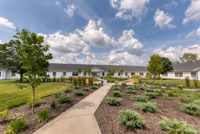 Wide exterior view of a single-story senior living facility building with a gray roof and white walls, surrounded by landscaped gardens with small bushes, trees, and a concrete walkway leading to the entrance under a partly cloudy blue sky.