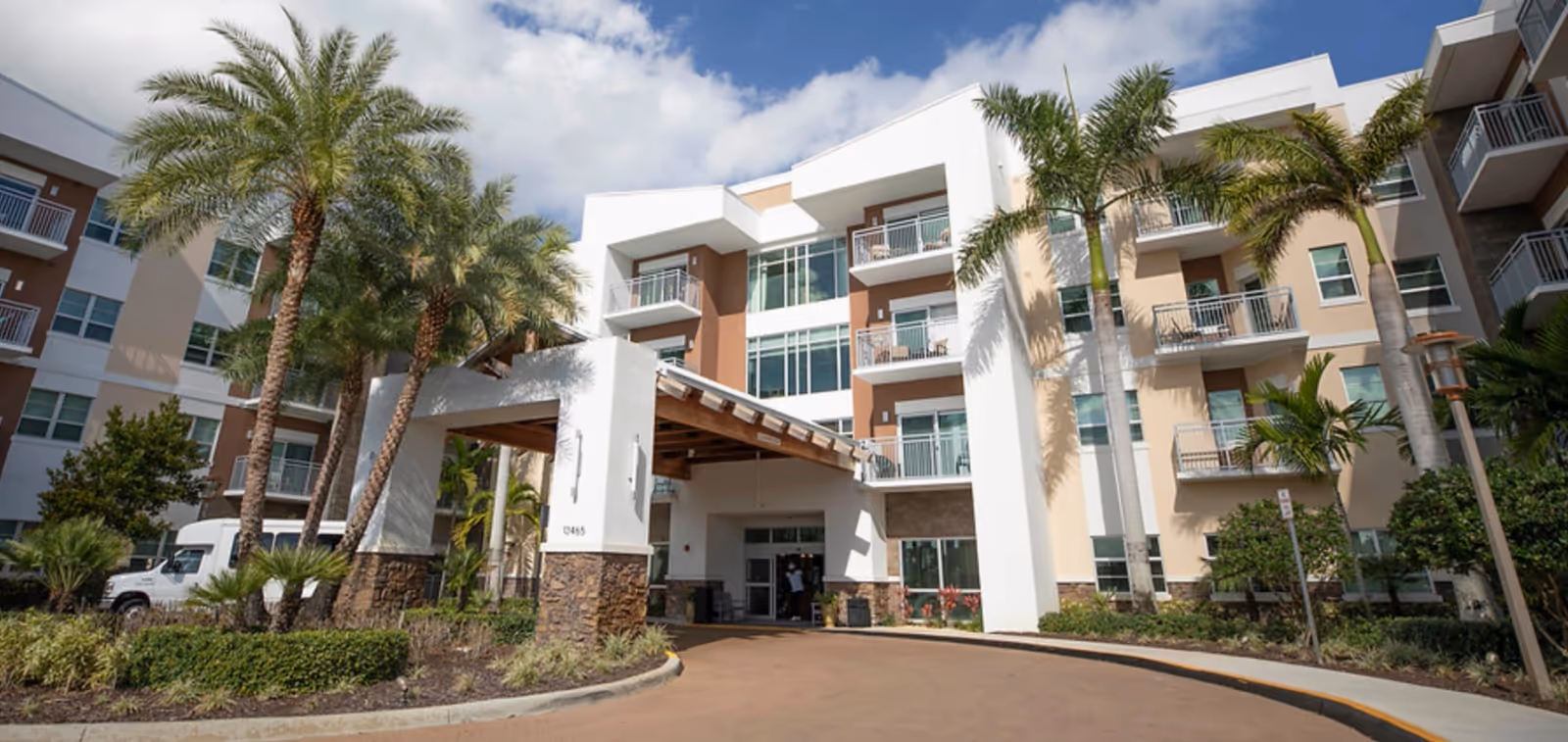 Front entrance of a multi-story senior living building with balconies, palm trees, and a covered porte-cochere.