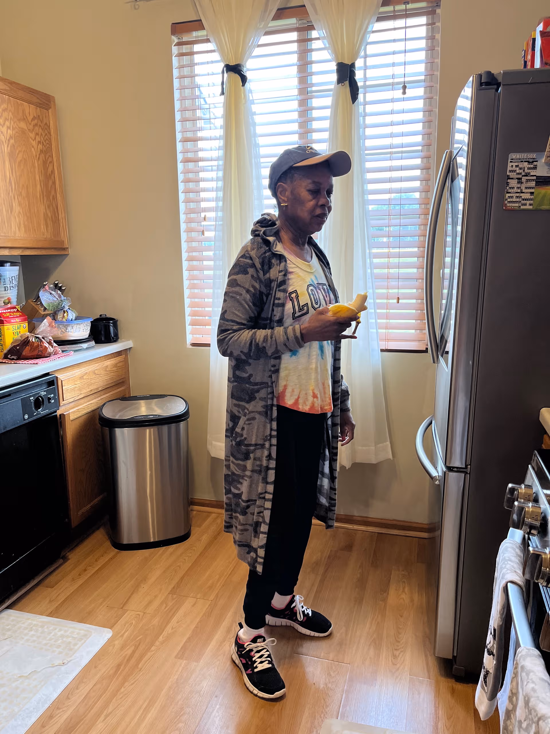 A senior woman stands in a bright kitchen beside a stainless refrigerator, holding a yellow object.