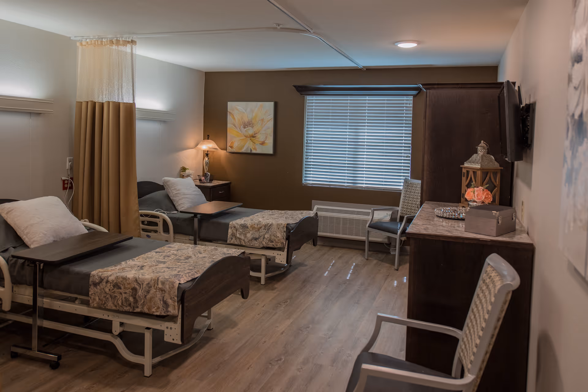 A rehabilitation and healthcare center room with two hospital beds, each with a pillow and patterned blanket. There is a curtain divider between the beds, a nightstand with a lamp and a flower, a window with blinds, two chairs, a dresser with decorative items, and a wall-mounted TV.