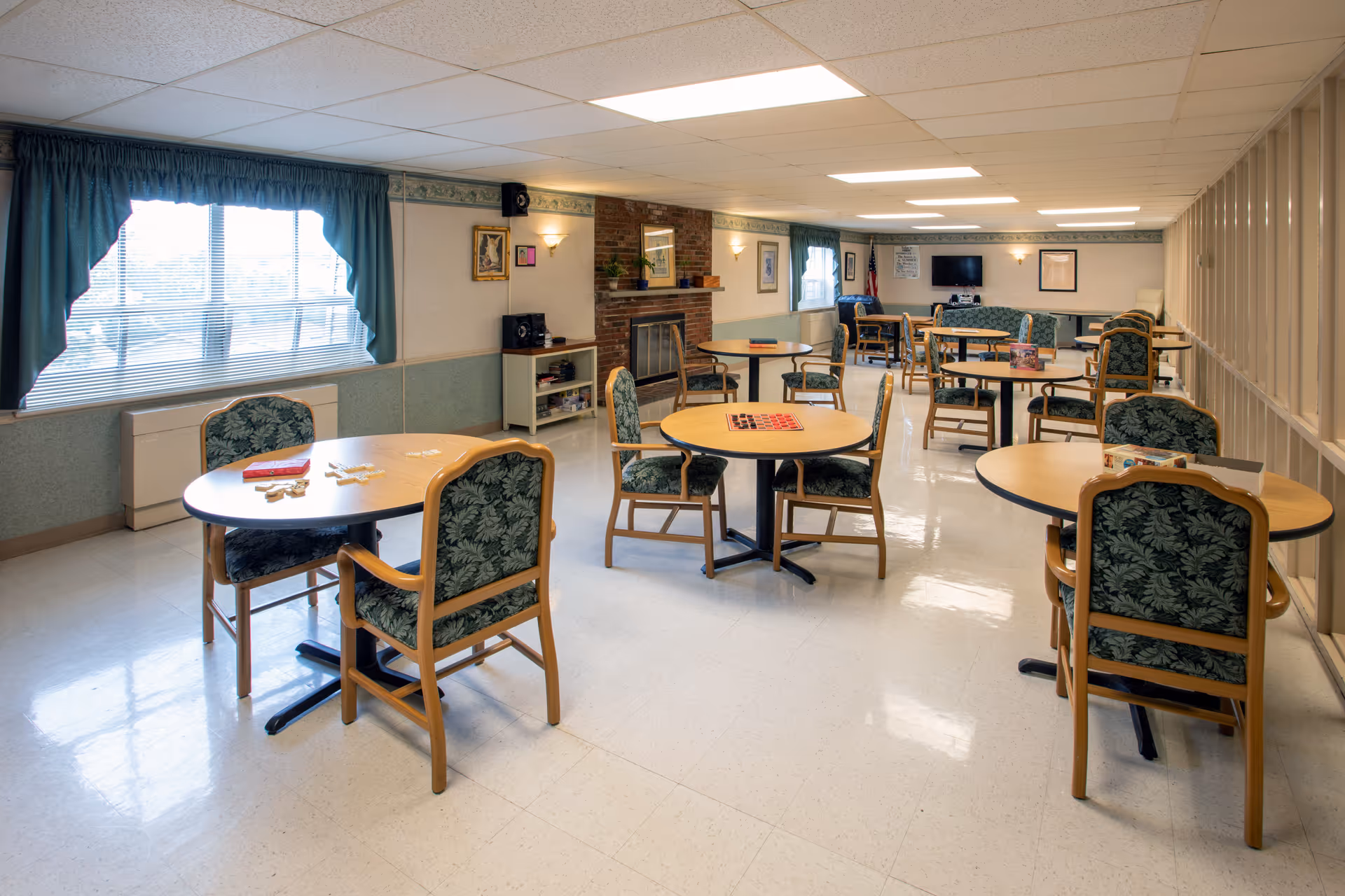 A spacious common room in a senior living facility with multiple round tables and chairs arranged for social activities. The room has large windows with teal curtains, a brick fireplace, framed pictures on the walls, and a television mounted on the far wall. Various board games and puzzles are placed on the tables.