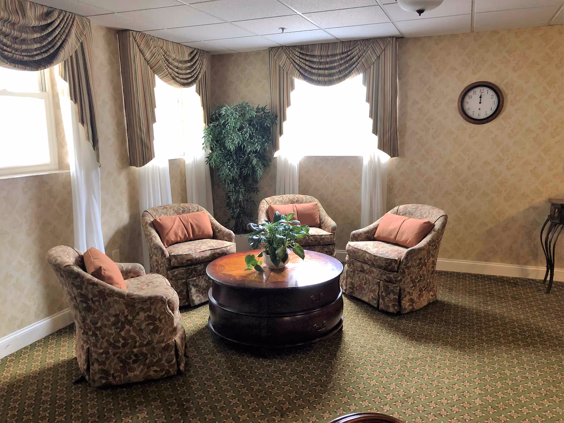Small sitting area with four upholstered armchairs around a round wooden coffee table beneath windows with draped curtains.
