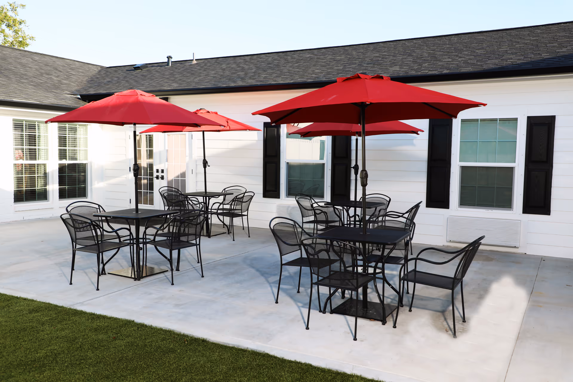 Outdoor patio area with black metal tables and chairs, each table shaded by a red umbrella. The patio is adjacent to a white building with black shutters and windows.
