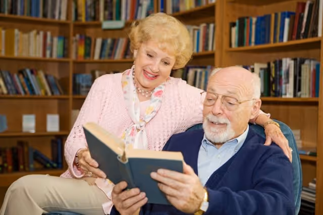 An elderly man and woman sitting together in a cozy room with bookshelves in the background. The man is seated in a chair reading a book, while the woman stands beside him, smiling and looking at the book over his shoulder.