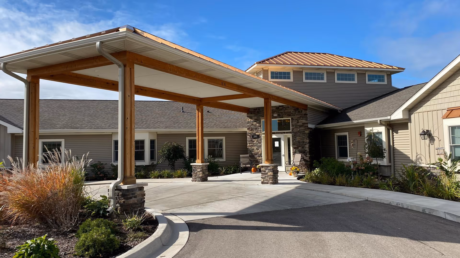 Covered porte-cochère at the front entrance of a single-story senior living building with stone accents, a metal roof, and landscaped planting beds.
