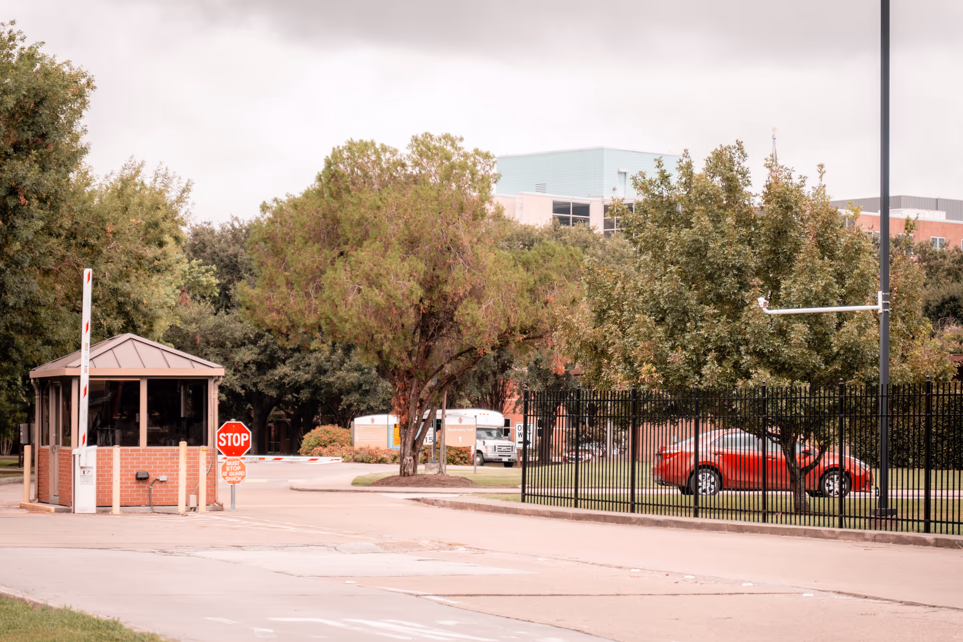 Entrance area with a small guardhouse, stop sign and barrier gate in front of a fenced building and trees.