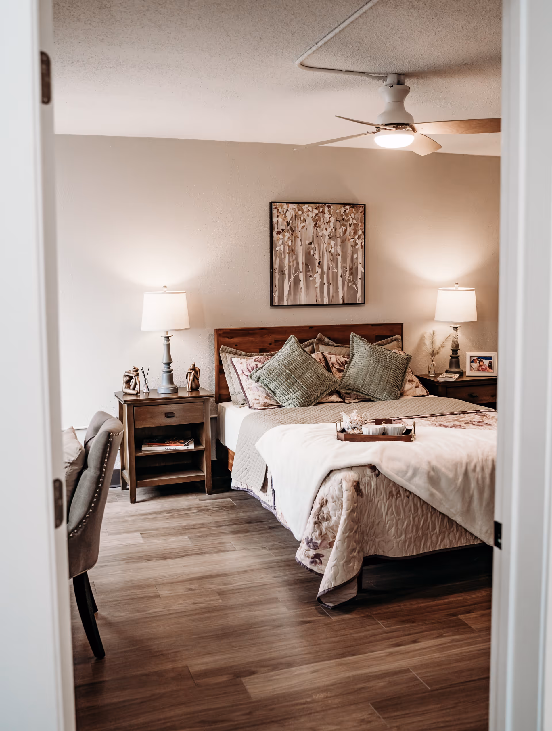Cozy furnished bedroom with a made bed, decorative pillows, bedside tables with lamps, wall art, and wood flooring.