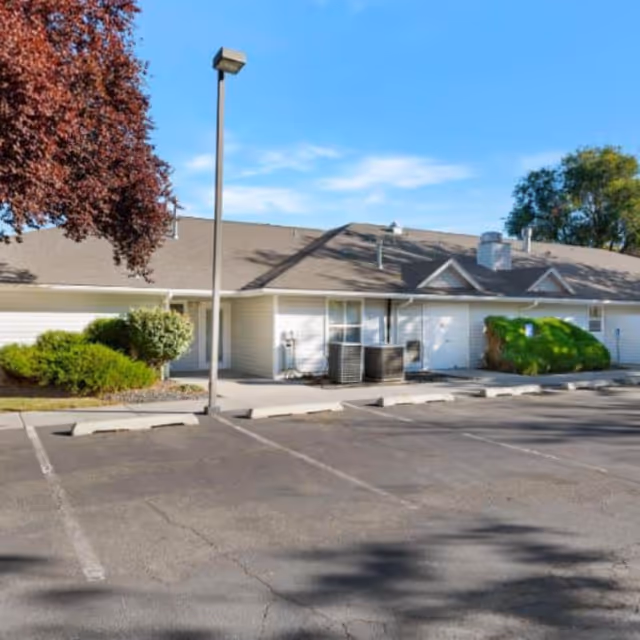 Single-story beige senior living facility exterior with a parking lot and surrounding shrubs.