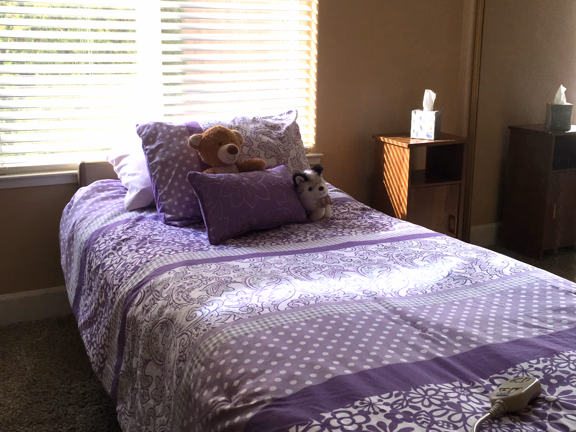 Purple patterned bed with stuffed animals in a sunlit bedroom.