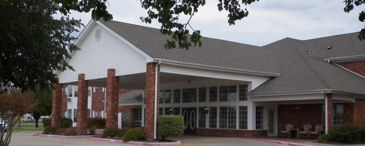Exterior view of a senior living facility building with a covered entrance supported by brick columns, large windows, and a seating area with chairs under a porch. Trees and landscaping surround the driveway.