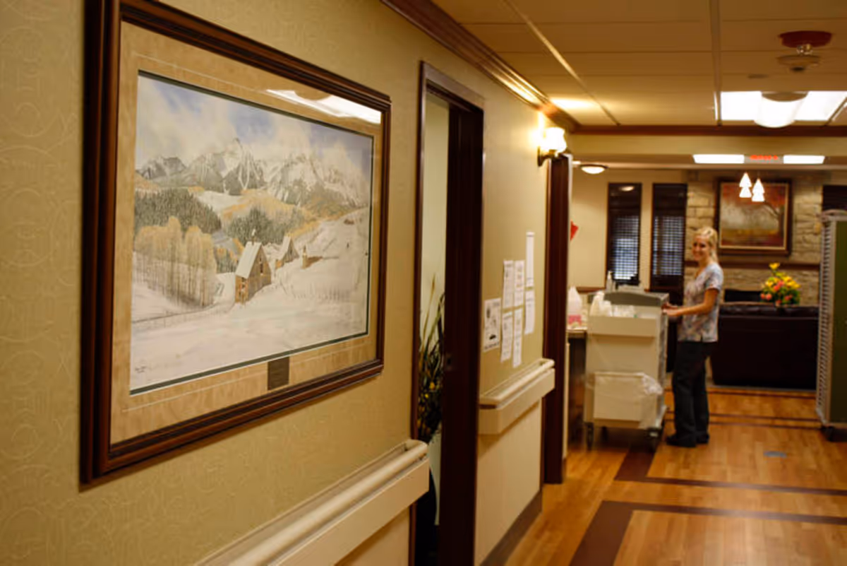 Interior hallway of St. Francis Manor showing a framed winter landscape on the wall and a staff member pushing a cart near a seating area.