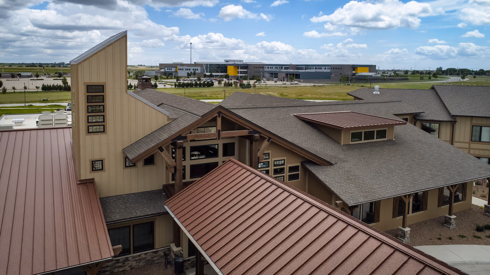 Aerial view of a senior living facility building with multiple roof sections, including brown and gray roofs, beige walls, and several windows. The building is surrounded by a paved walkway and landscaping with small plants. In the background, there is a large modern building and open grassy fields under a partly cloudy sky.