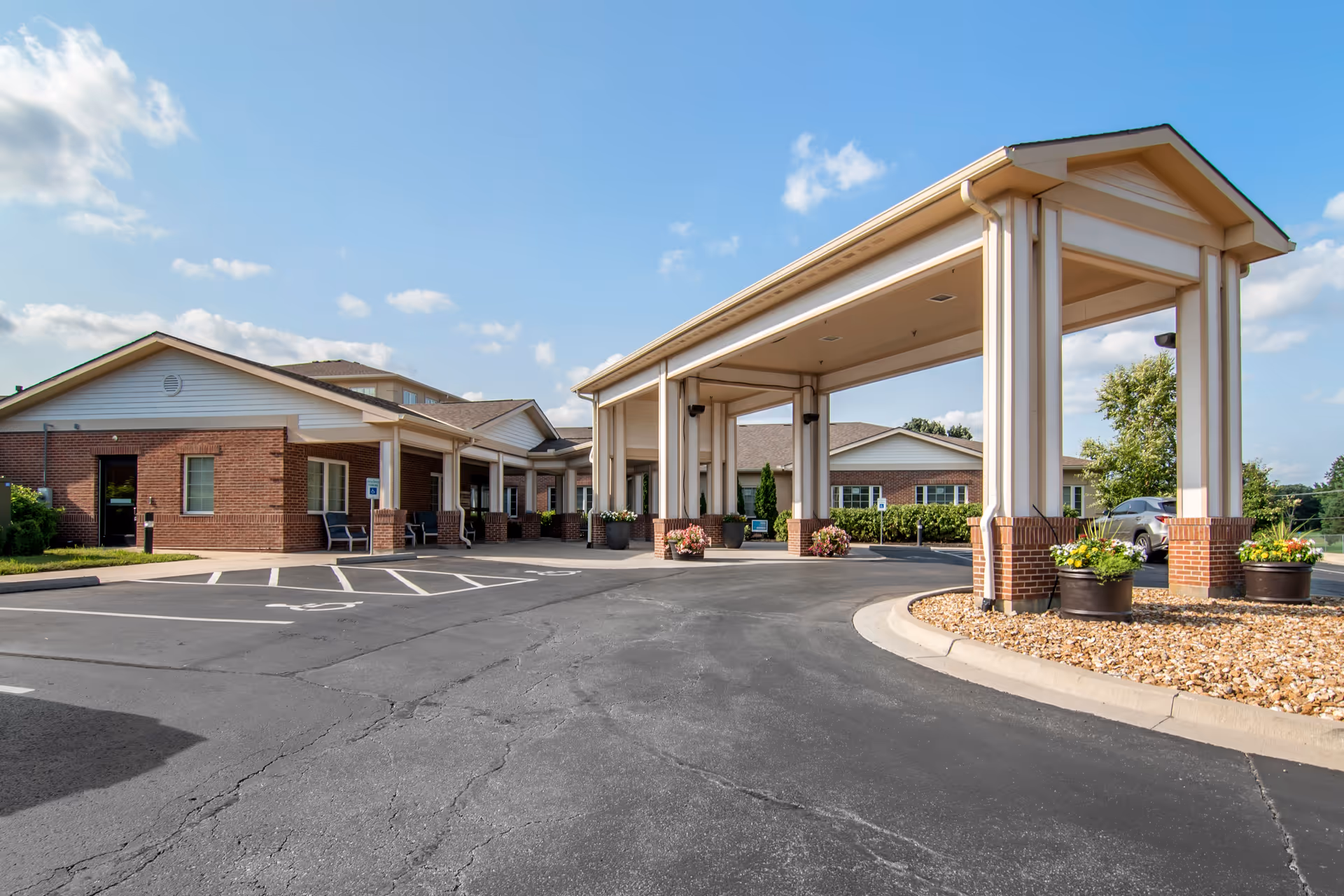 Exterior view of Stratford Commons Rehabilitation & Health Care facility showing the entrance with a covered drop-off area, brick and siding building, parking spaces including a handicapped spot, and landscaped areas with potted plants under a partly cloudy sky.