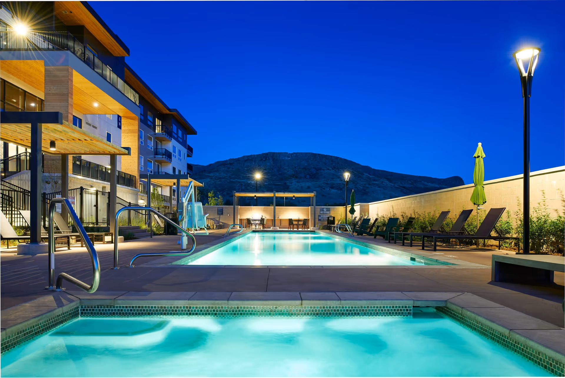 Lit outdoor swimming pool and adjacent hot tub at dusk beside a modern multi-story residential building with lounge chairs and umbrellas.