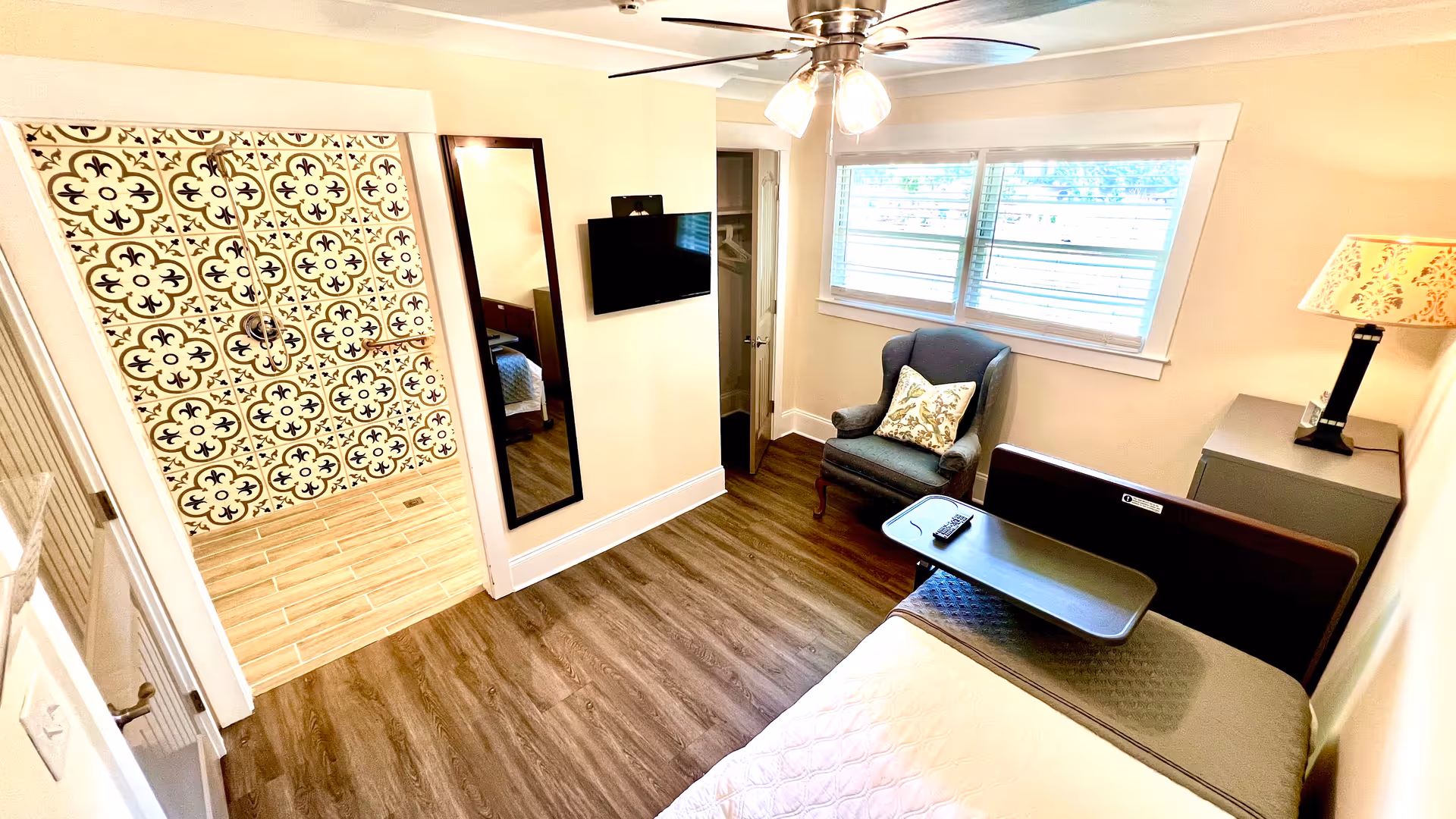 A bright senior living bedroom with wood flooring, a bed with an overbed table, a comfortable armchair with a decorative pillow, a wall-mounted TV, a full-length mirror, a bedside table with a lamp, and a walk-in shower with patterned tile walls.