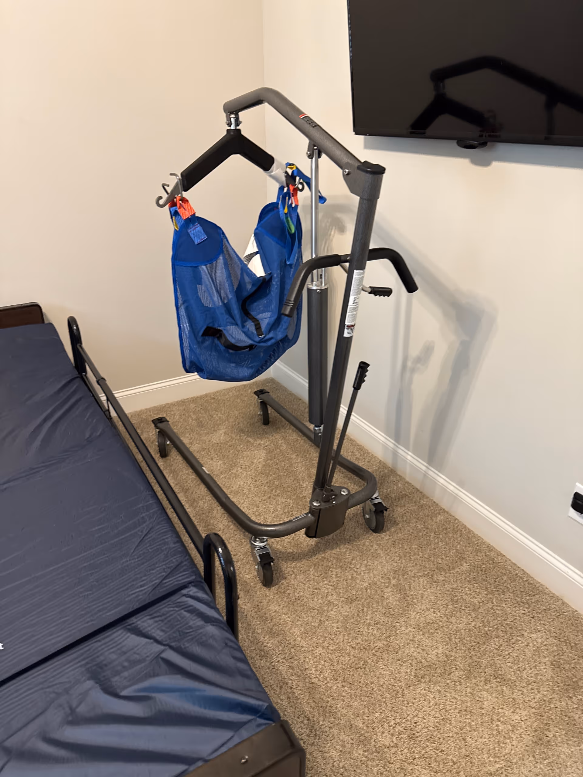 A blue patient lift sling hangs from a stand beside a bed in a small bedroom.