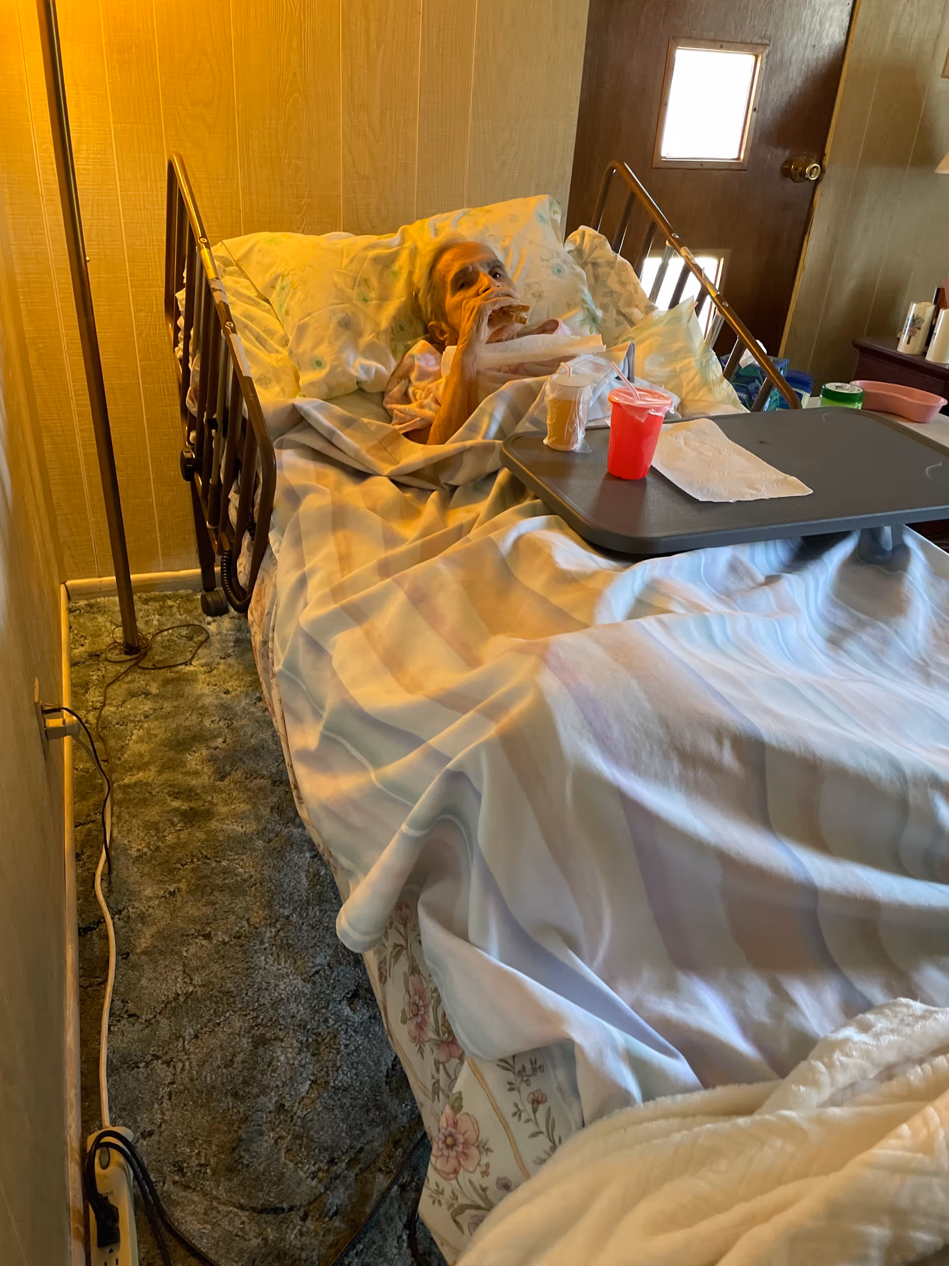 Elderly patient lies in a hospital-style bed with a tray table in a private room.