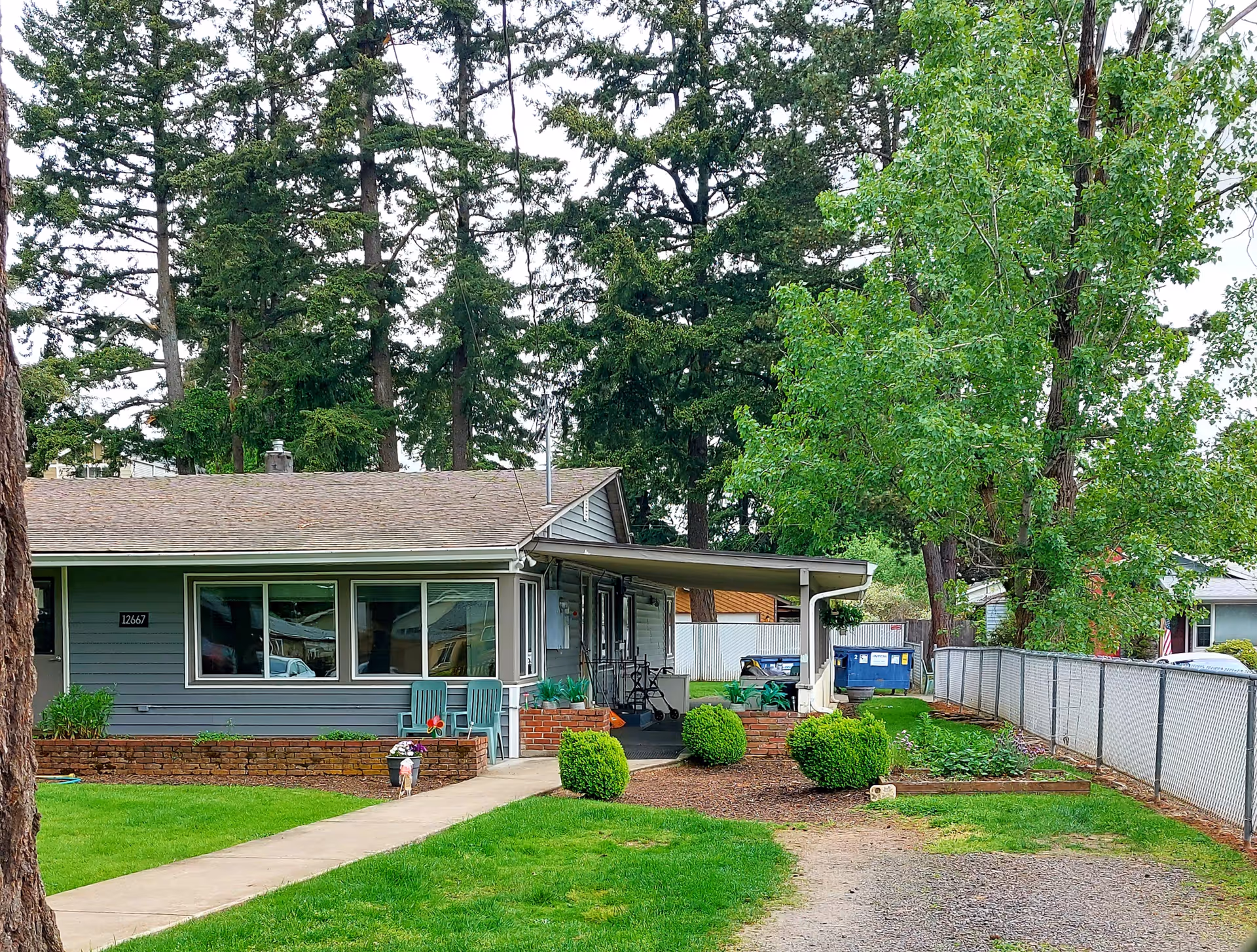 Single-story gray house with front porch, manicured lawn, and tall trees behind.