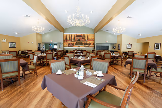 A spacious dining room with multiple tables covered in brown tablecloths, each set with white cups, glasses, and napkins. The room features wooden flooring, green cushioned chairs, chandeliers hanging from the ceiling, and a large mural on the far wall depicting a countryside scene with trees and a building.