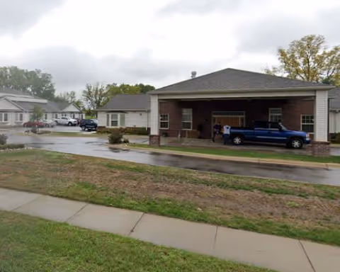 Exterior view of a brick senior living facility with a covered entry and parked vehicles.