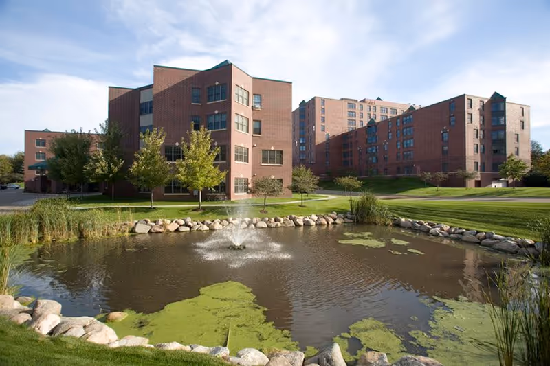 Brick multi-story senior living building set behind a landscaped lawn and a pond with a small fountain.