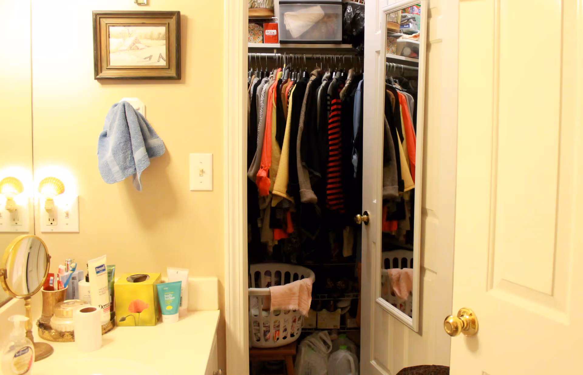 A bathroom vanity area with a countertop holding various toiletries, a small round mirror, a roll of toilet paper, and a tissue box. A blue towel hangs on a hook on the wall above the light switch. To the right, there is an open door revealing a closet filled with hanging clothes and a laundry basket. The closet door has a full-length mirror attached.